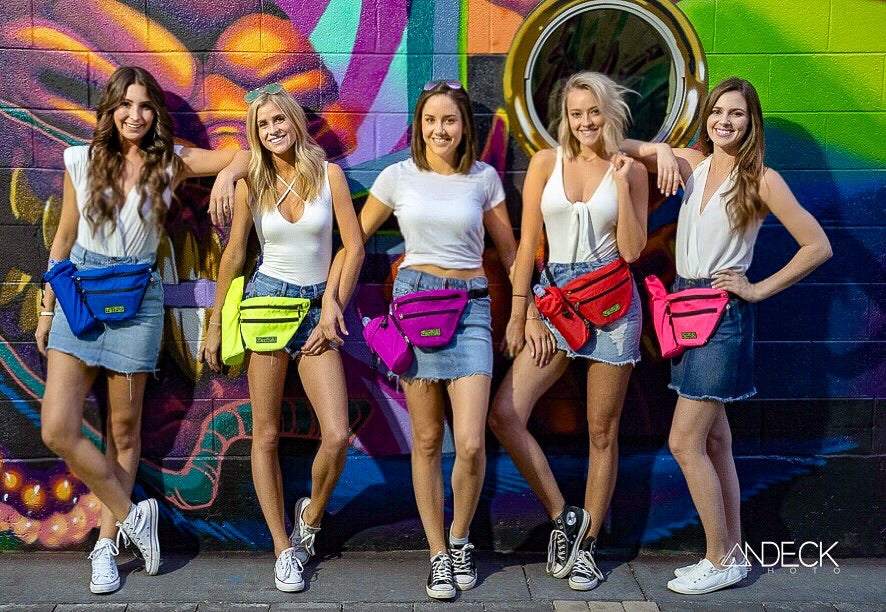 Group of five women wearing colorful FezPak customizable fanny packs with attachable bottle holders against a vibrant graffiti wall