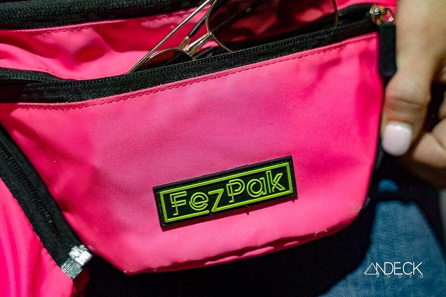 Bright pink FezPak customizable fanny pack with black zippers and logo patch, shown holding sunglasses inside pocket.