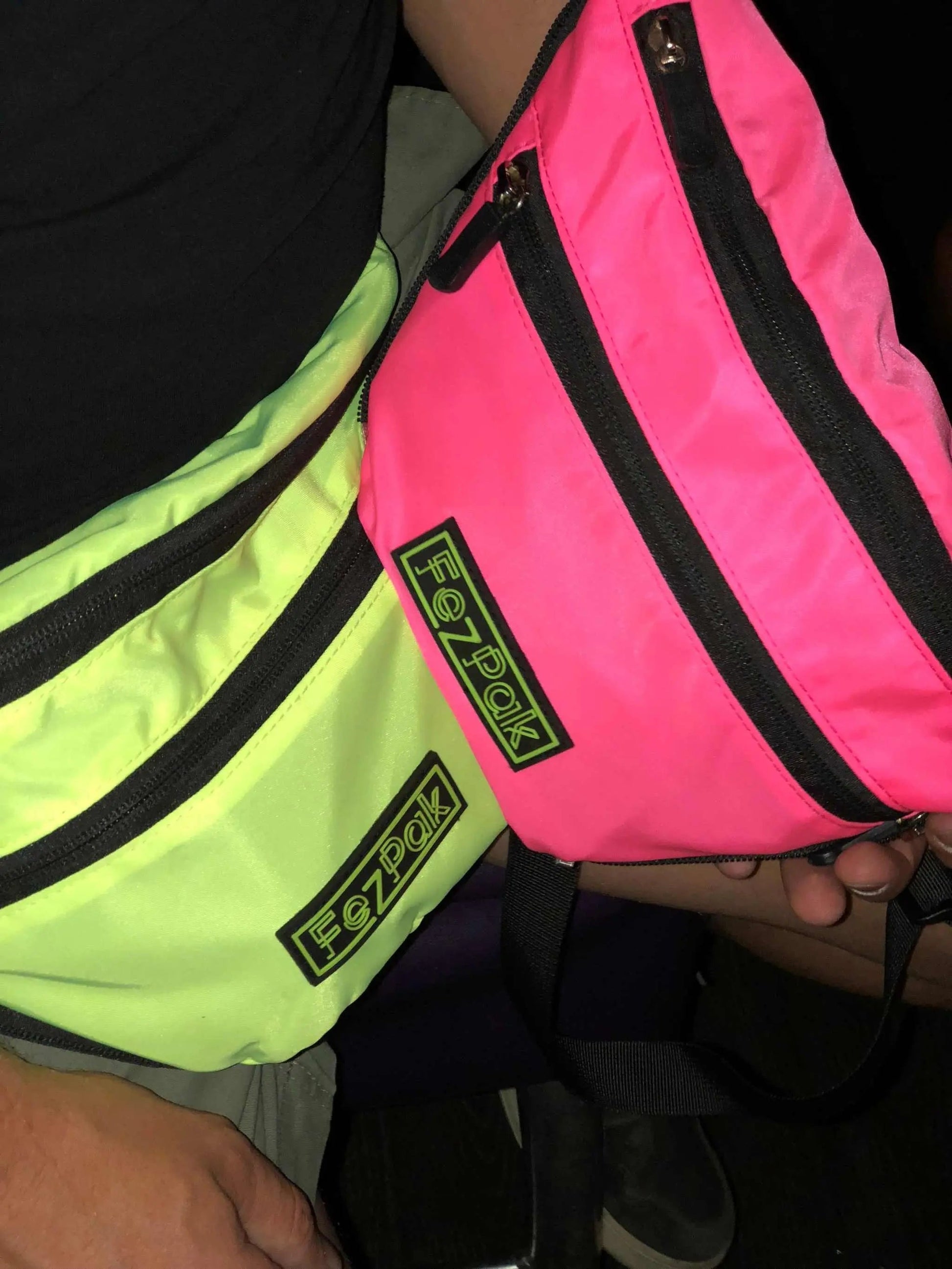 Two FezPak customizable flat design fanny packs in neon yellow and pink with black zippers and logo patches worn crossbody and waist style.